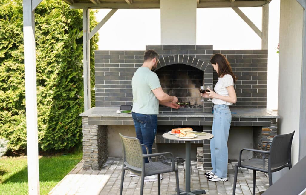 A man grilling meat at an outdoor kitchen with a woman holding a glass of wine nearby. The setting features a stone countertop, a brick grill, and a table with food items, surrounded by greenery and a paved patio.