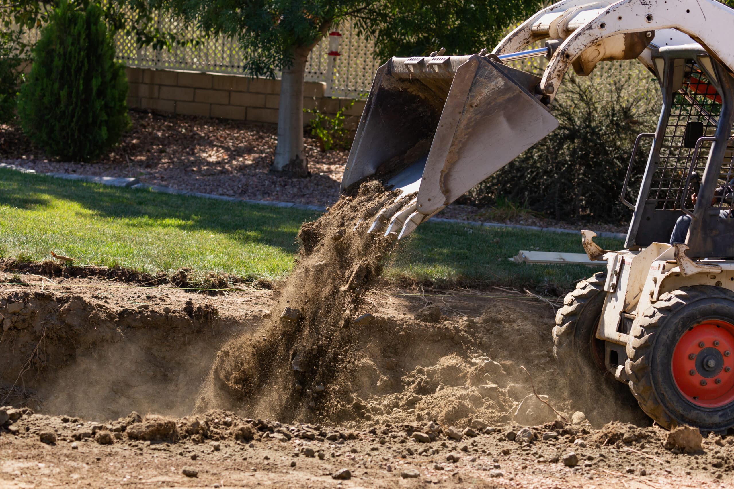A backhoe loader is raising its bucket filled with freshly dug soil, releasing a cloud of dust. The surrounding area features green grass, shrubs, and a fence in the background, indicating an outdoor construction or landscaping project.