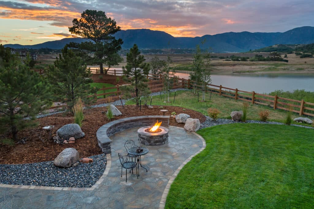 An outdoor landscape featuring a stone patio with a fire pit surrounded by rocks and chairs. In the background, trees and a fence are visible, along with a lake and mountains under a sunset sky with clouds.
