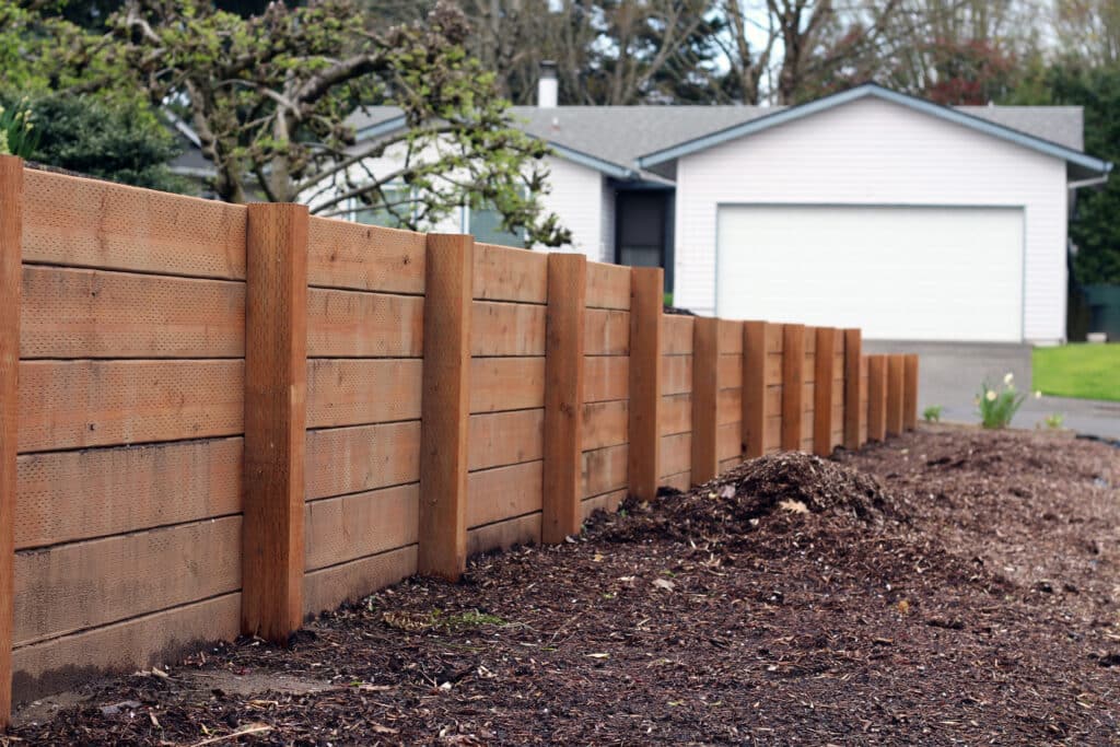 A wooden fence made of horizontal planks, extending along the edge of a landscaped area. In the background, a single-story house with a garage door is visible, set against a backdrop of trees and shrubbery.